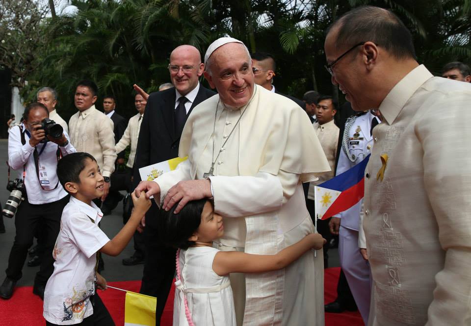 Pope Francis is being hugged by one child while another clutches his hand. He is surrounded by papal aides and Filipino officials as he arrives in the Philippines outdoors.